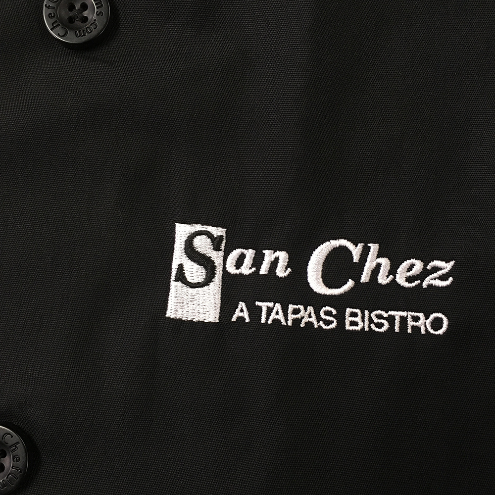 White and black San Chez A Tapas Bistro embroidery on a black chef's coat.