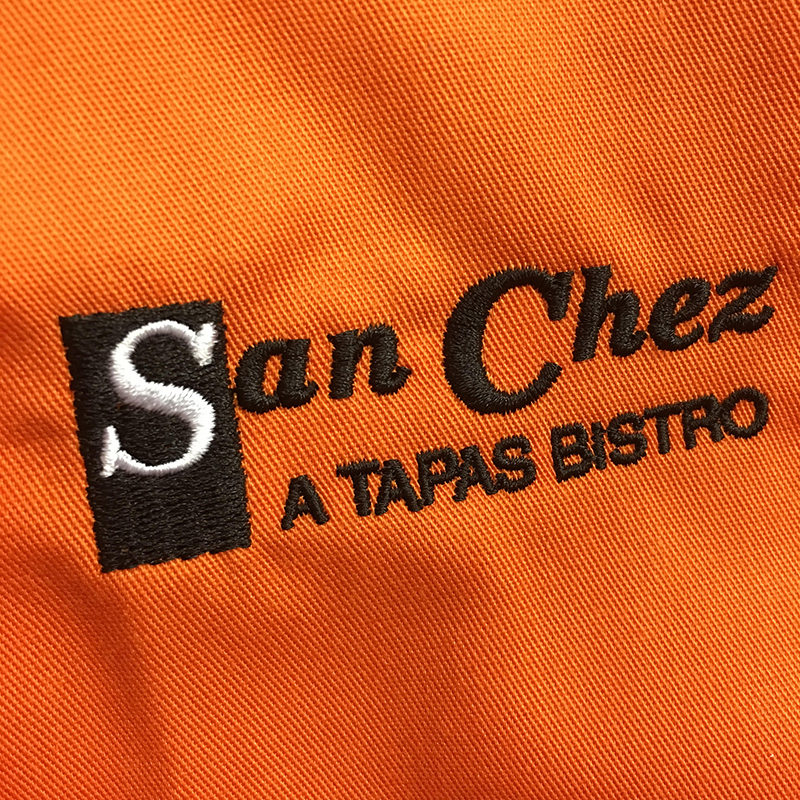 Black and White San Chez A Tapas Bistro Embroidery on bright orange chef's coat.